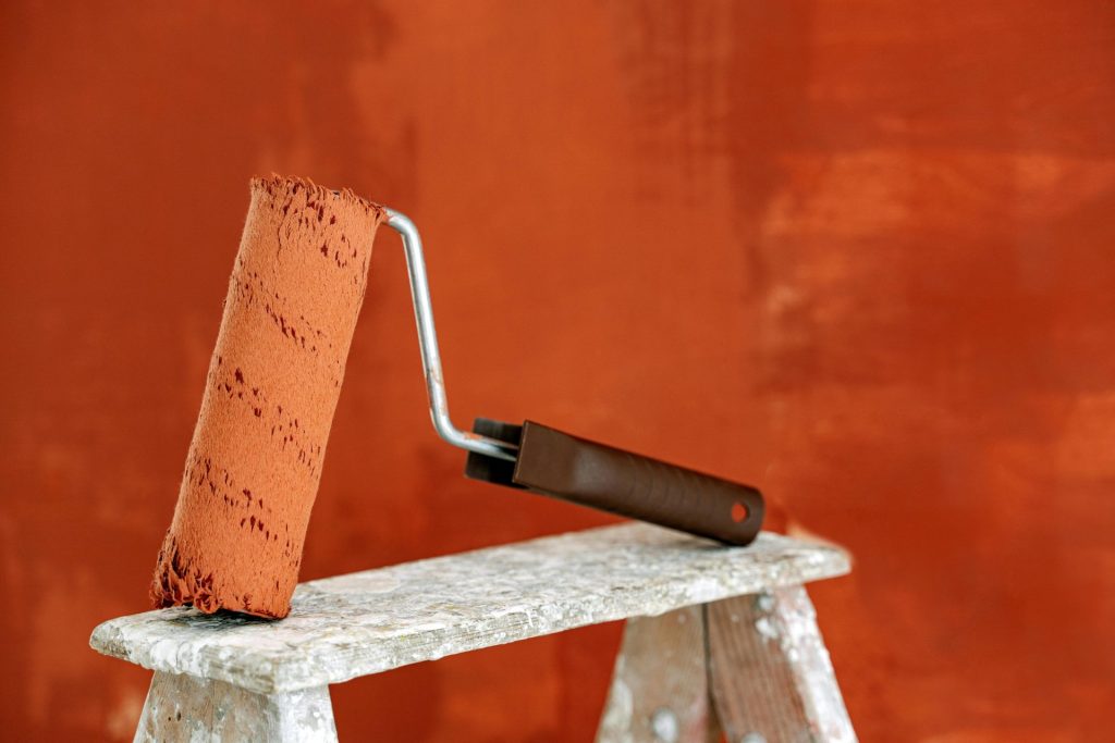 Detailaufnahme einer Farbrolle, die auf einer Leiter ruht; im Hintergrund ist eine leuchtend orangefarbene Wand zu sehen.